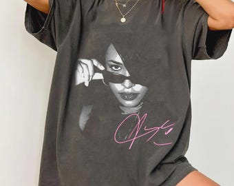 Puede incluir: Camiseta gris oscuro con una imagen en blanco y negro de una persona con gafas de sol. La imagen es un primer plano de la cara de la persona. La camiseta también tiene una firma rosa en la parte delantera.