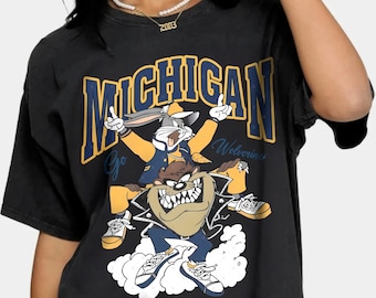 Puede incluir: Camiseta negra con un gráfico de Bugs Bunny y Taz de Looney Tunes. El gráfico incluye el texto "Michigan" y "Go Wolverines".