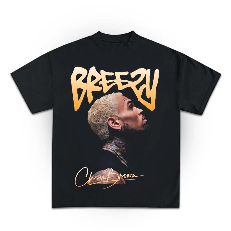 May include: Black t-shirt with a profile image of a person with blonde hair and tattoos. The word "BREEZY" is printed in an orange gradient font above the image. The signature "Chris Brown" is printed in gold below the image.