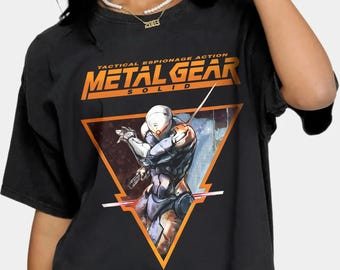 Puede incluir: Camiseta negra con un gráfico de un personaje de la serie de videojuegos Metal Gear Solid. El gráfico está en forma de triángulo con el texto "Tactical Espionage Action Metal Gear Solid" arriba.