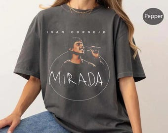 May include: Dark gray t-shirt featuring a singer with a microphone inside a white circle, with the word "MIRADA" in a handwritten font. The text "IVAN CORNEJO" is above the image. The shirt is a Pepper color.