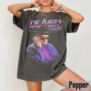 May include: A charcoal gray oversized t-shirt featuring a graphic print of Oliver Tree wearing red sunglasses and a purple jacket. The text OLIVER TREE is printed in a purple and white gradient font above the portrait. The shirt has short sleeves.
