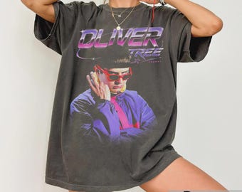 Puede incluir: Una camiseta gris carbón de corte oversize con un estampado gráfico de Oliver Tree con gafas de sol rojas y una chaqueta morada. El texto OLIVER TREE aparece impreso en un degradado morado y blanco sobre el retrato. Manga corta.