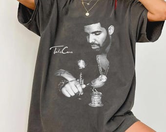 Puede incluir: Camiseta gris oscuro con una imagen en blanco y negro de un hombre y el texto "Take Care". El gráfico muestra al hombre sosteniendo un pájaro sobre un pedestal. La camiseta tiene mangas cortas y un corte holgado.