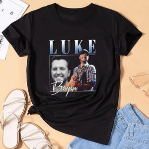 May include: Black t-shirt featuring a portrait of Luke Bryan with the name "LUKE" in blue and white block letters. The word "Bryan" is written in a cursive font below the portrait. The shirt is paired with sandals and denim shorts.