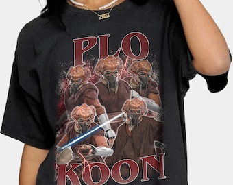 Puede incluir: Camiseta negra con un gráfico de Plo Koon de Star Wars. El diseño incluye múltiples imágenes del personaje, con el nombre "PLO KOON" en una fuente roja desgastada. El personaje se representa con una máscara y un sable de luz.