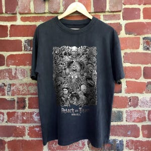 May include: Black t-shirt with a white graphic print of characters from the anime Attack on Titan. The text "Attack on Titan" and "進撃の巨人" are printed below the graphic.