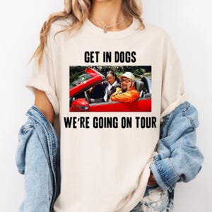 May include: A cream-coloured t-shirt with the text "GET IN DOGS WE'RE GOING ON TOUR" in black. The shirt features a graphic of two people in a red convertible. The person in the passenger seat is wearing an orange jacket and a hat.