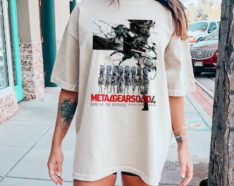 Puede incluir: Una camiseta blanca con un estampado gráfico de un personaje de videojuego y el texto "Metal Gear Solid 4: Guns of the Patriots Tactical Espionage Action".