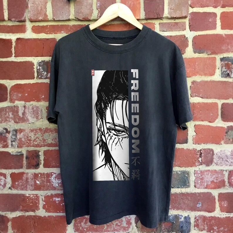 May include: Dark grey t-shirt with a black and white illustration of a character's face. The word "FREEDOM" is printed vertically in grey on the right, with Japanese characters below. The shirt is displayed on a wooden hanger.