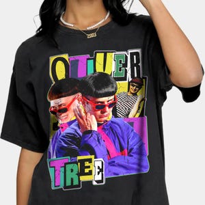 Pode incluir: Camiseta preta com um gráfico colorido de Oliver Tree. O gráfico apresenta um retrato de Oliver Tree com o texto "Oliver Tree" em uma fonte retrô colorida.
