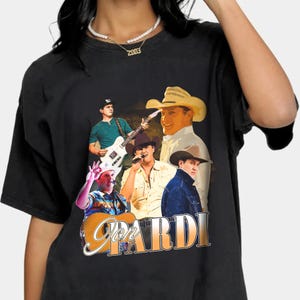 Puede incluir: Camiseta negra con un collage del cantante de música country, Jon Pardi, con un sombrero de vaquero y sosteniendo una guitarra. El texto "Jon Pardi" está impreso en letras doradas en la camiseta.