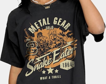 Puede incluir: Camiseta negra con un gráfico de estilo vintage de dos personajes montando una motocicleta. El gráfico incluye el texto "Metal Gear Solid Snake Eater 1964 What a Thrill".