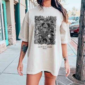 May include: A beige oversized t-shirt with a black and white graphic print of characters from the anime "Attack on Titan". The graphic features a large, menacing titan with other characters surrounding it. The text "Attack on Titan" and "Shingeki no Kyojin" are printed below the graphic.