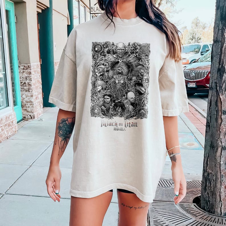 May include: A beige oversized t-shirt with a black and white graphic print of characters from the anime "Attack on Titan". The graphic features a large, menacing titan with other characters surrounding it. The text "Attack on Titan" and "Shingeki no Kyojin" are printed below the graphic.