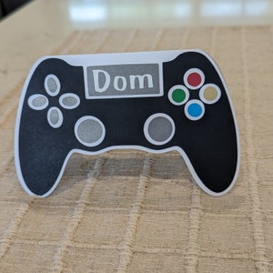 Op de afbeelding: Zwart-witte gamecontroller met een wit label met de tekst "Dom" en gekleurde knoppen.