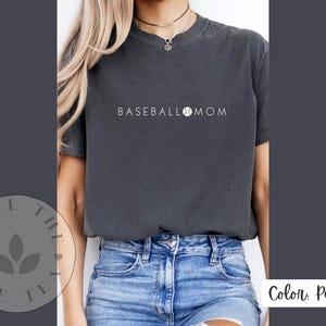 May include: A dark grey t-shirt with the text "BASEBALL MOM" and a baseball graphic. The shirt is being worn by a person with long brown hair and blue jeans.