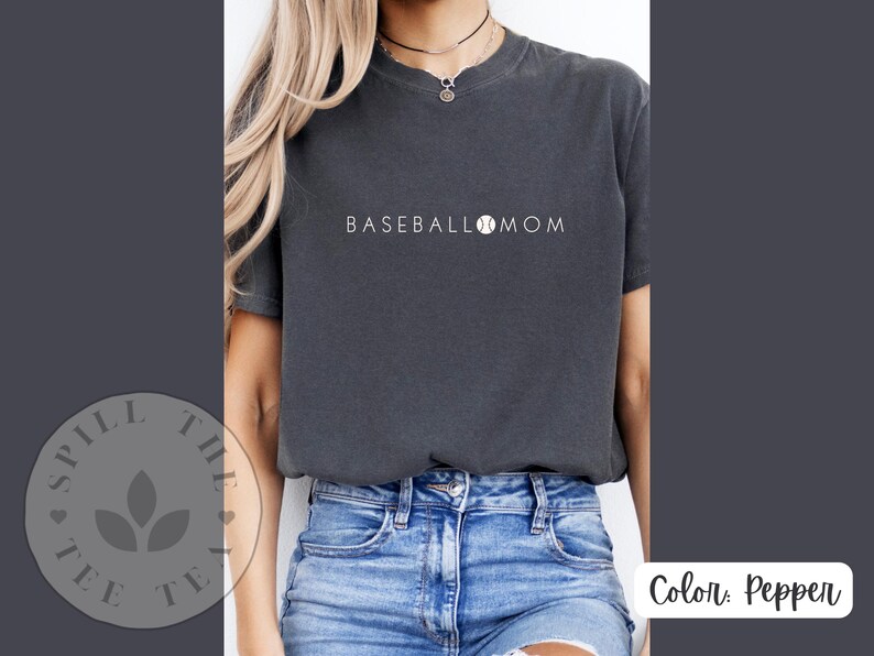 May include: A dark grey t-shirt with the text "BASEBALL MOM" and a baseball graphic. The shirt is being worn by a person with long brown hair and blue jeans.