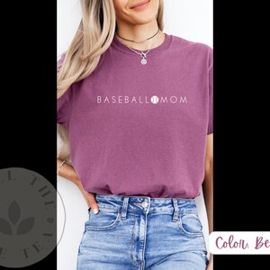 May include: A woman wearing a purple t-shirt with the text "BASEBALL MOM" and a baseball graphic. She is wearing blue jeans.