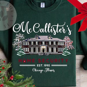 Puede incluir: Una sudadera de cuello redondo verde oscuro con un gr&aacute;fico blanco de una casa grande y &aacute;rboles nevados. El texto dice "McCallister's HOME SECURITY EST. 1990 Chicago, Illinois" en letras blancas y rojas, con un lazo rojo en la parte superior derecha.