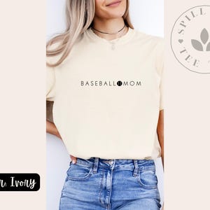 May include: Ivory t-shirt with the text "BASEBALL MOM" and a baseball graphic. The shirt is being modelled by a person wearing blue jeans. The brand name "Spill the Tee Tea" is visible in a circle with a leaf design.