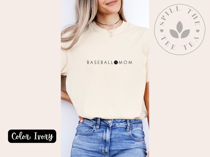 May include: Ivory t-shirt with the text "BASEBALL MOM" and a baseball graphic. The shirt is being modelled by a person wearing blue jeans. The brand name "Spill the Tee Tea" is visible in a circle with a leaf design.