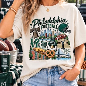 Könnte beinhalten: Ein cremefarbenes T-Shirt mit einem Grafikdruck von Wahrzeichen Philadelphias, einem Football-Helm, einem Football, einem Cheesesteak, einer Brezel, einem Adler mit Sonnenbrille und Mütze sowie dem Text Philadelphia Football und City of Brotherly Love.