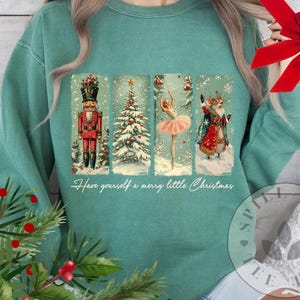 Puede incluir: Sudadera de color verde azulado con un diseño navideño festivo. El gráfico incluye un cascanueces, un árbol de Navidad, una bailarina y un ratón en una escena invernal. El texto "Have yourself a merry little Christmas" está impreso debajo.