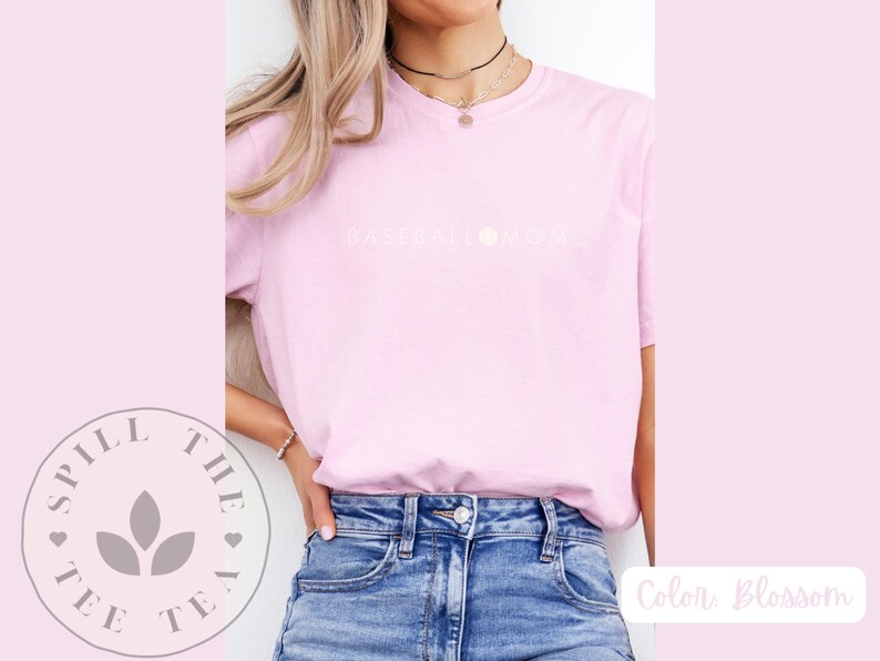 May include: A pink t-shirt with the text "Baseball Mom" in white lettering. The shirt is being worn by a person with long blonde hair. The person is wearing blue jeans.