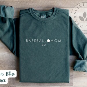 May include: A dark green sweatshirt with the text "BASEBALL MOM #2" in white. The sweatshirt features a baseball graphic and is made from a soft, comfortable fabric.