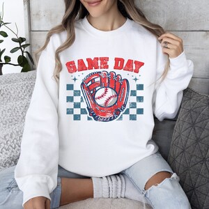 Pu&ograve; includere: Felpa bianca con una grafica di guanto da baseball e palla rosso, bianco e blu. Il testo "GAME DAY" &egrave; stampato in blu sopra la grafica.