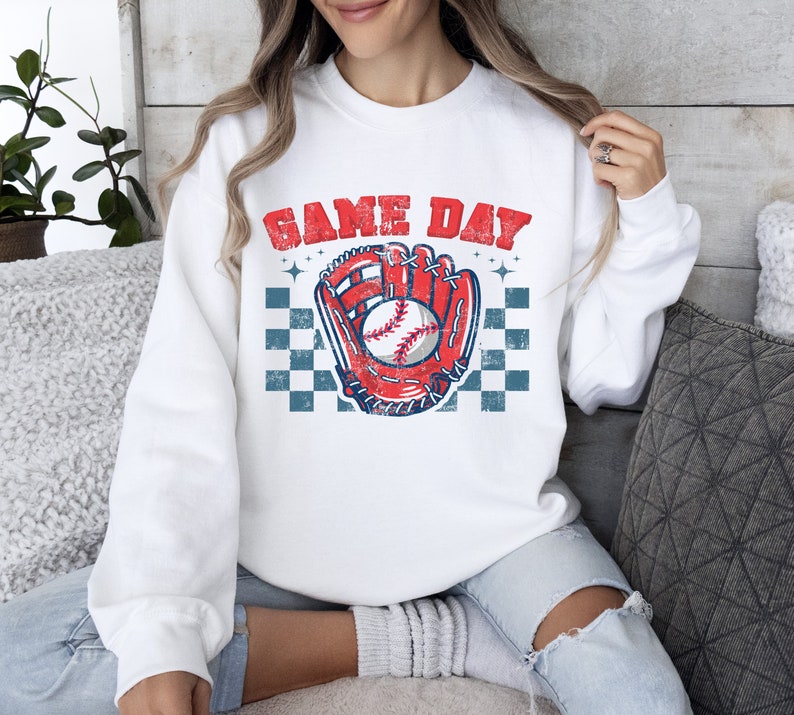 Pu&ograve; includere: Felpa bianca con una grafica di guanto da baseball e palla rosso, bianco e blu. Il testo "GAME DAY" &egrave; stampato in blu sopra la grafica.
