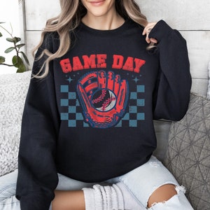 Pu&ograve; includere: Felpa nera con una grafica di guanto da baseball rosso e blu e una palla. Il testo "GAME DAY" &egrave; stampato in bianco sopra la grafica.