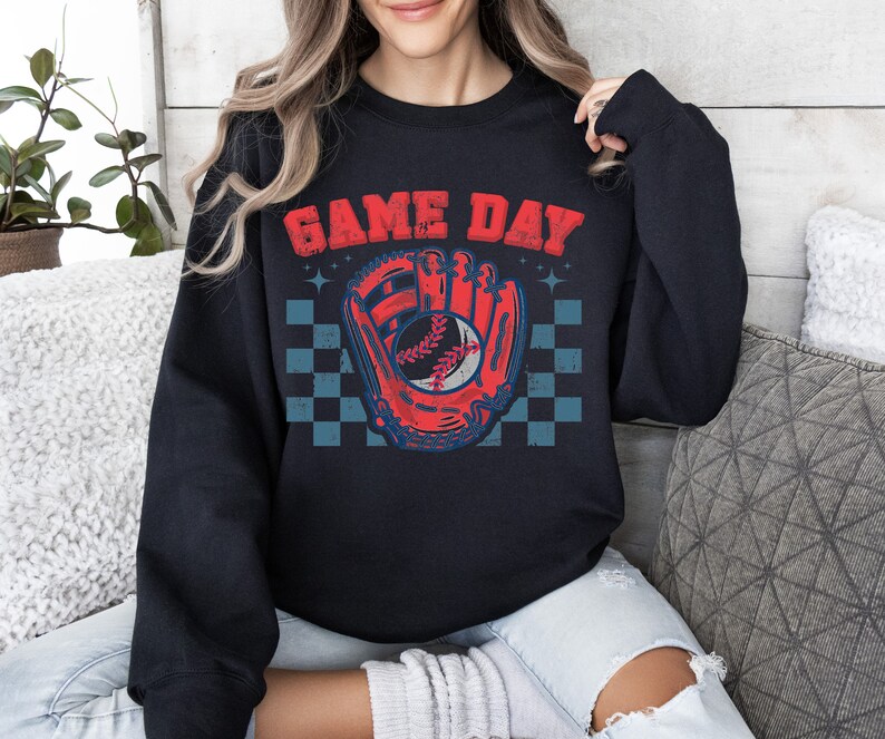 Pu&ograve; includere: Felpa nera con una grafica di guanto da baseball rosso e blu e una palla. Il testo "GAME DAY" &egrave; stampato in bianco sopra la grafica.