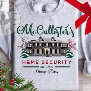 Puede incluir: Una sudadera gris claro de cuello redondo con un gr&aacute;fico de una casa grande y &aacute;rboles nevados. El texto dice: McCallister's HOME SECURITY EST. 1990 Chicago, Illinois. El dise&ntilde;o incluye un icono de candado rojo y letras de color verde oscuro.