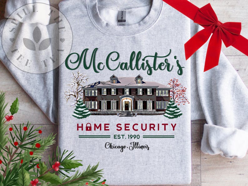 Puede incluir: Una sudadera gris claro de cuello redondo con un gr&aacute;fico de una casa grande y &aacute;rboles nevados. El texto dice: McCallister's HOME SECURITY EST. 1990 Chicago, Illinois. El dise&ntilde;o incluye un icono de candado rojo y letras de color verde oscuro.