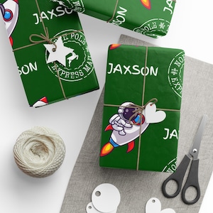 May include: Green gift wrap with the name "Jaxson" in white, decorated with a rocket ship and an astronaut. The gifts are tied with twine and adorned with star and heart-shaped tags. A spool of twine and scissors are also visible.