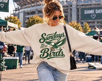 May include: A cream-colored crewneck sweatshirt featuring a dark green graphic print that reads "Sundays ARE FOR THE Birds" with an illustration of an eagle holding a football, accented by small stars and a heart, worn with blue denim jeans.