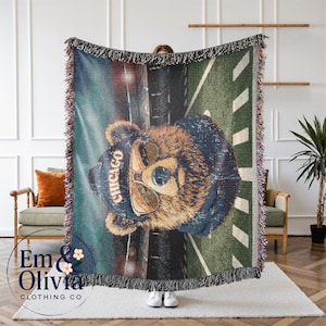 May include: A woven throw blanket featuring a bear wearing sunglasses and a Chicago hat. The blanket has a green and blue background with a fringe border. The bear is the central focus of the design.