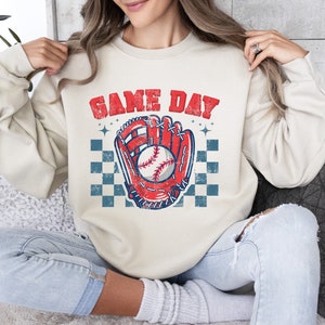 Pu&ograve; includere: Una felpa color crema con il testo "GAME DAY" in lettere rosse e blu. Un guanto da baseball rosso con una palla da baseball bianca &egrave; al centro della felpa. Il guanto &egrave; su uno sfondo a scacchi blu e bianco.