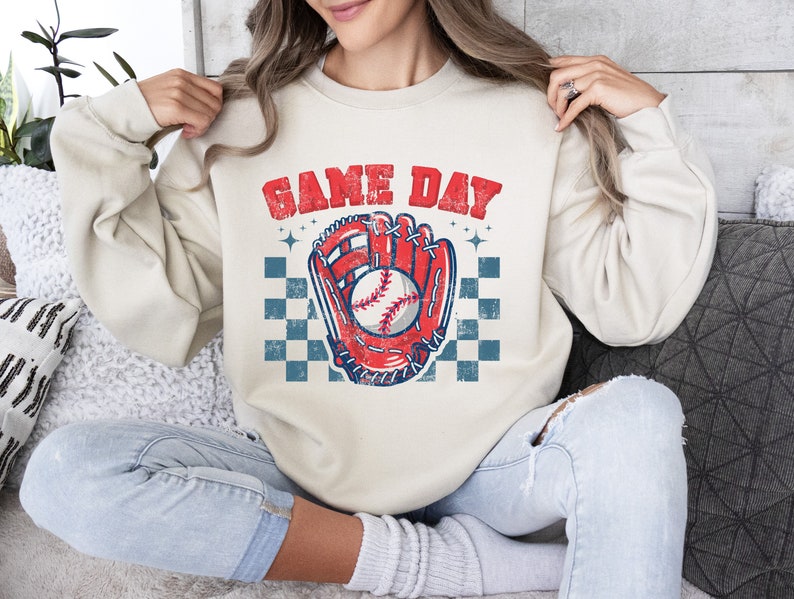 Pu&ograve; includere: Una felpa color crema con il testo "GAME DAY" in lettere rosse e blu. Un guanto da baseball rosso con una palla da baseball bianca &egrave; al centro della felpa. Il guanto &egrave; su uno sfondo a scacchi blu e bianco.