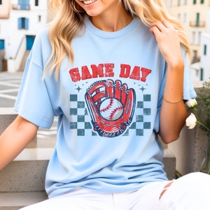 Pu&ograve; includere: Una maglietta blu chiaro con una grafica di baseball vintage e il testo "GAME DAY". La grafica presenta un guanto da baseball rosso con una palla da baseball bianca al suo interno.