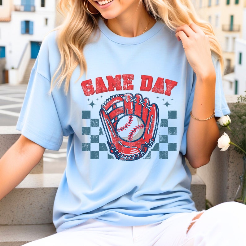 Pu&ograve; includere: Una maglietta blu chiaro con una grafica di baseball vintage e il testo "GAME DAY". La grafica presenta un guanto da baseball rosso con una palla da baseball bianca al suo interno.