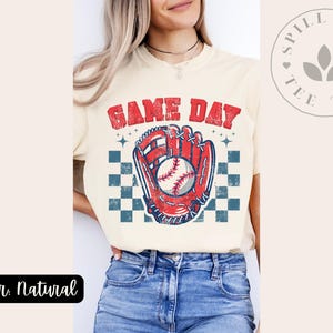 Pu&ograve; includere: Una maglietta color crema con una grafica di baseball vintage e il testo "GAME DAY".