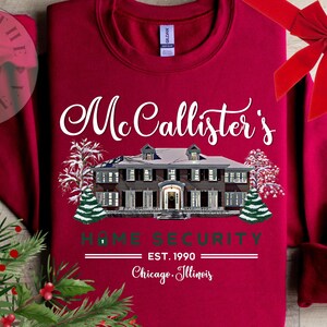 Puede incluir: Una sudadera de cuello redondo color rojo oscuro con el gr&aacute;fico de una casa grande cubierta de nieve. El texto dice McCallister's HOME SECURITY EST. 1990 Chicago, Illinois. El dise&ntilde;o incluye &aacute;rboles nevados y un icono de candado.