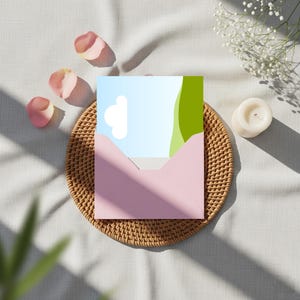 Könnte beinhalten: Ein rosa Umschlag enthält eine Karte mit einem blauen Himmel und einer grünen Landschaft. Die Szene ist auf einem geflochtenen Untersetzer angeordnet, mit Rosenblättern und einer brennenden Kerze. Weiche Schatten verstärken die Komposition.