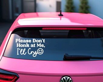 May include: The rear window of a pink car features a white decal with the words "Please Don't Honk at Me, I'll Cry" and a smiley face. The car has a black aerial and a rear wiper.