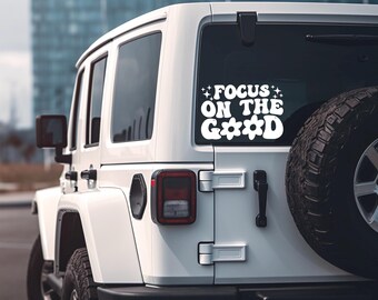 May include: White SUV with a spare tire on the back. A white decal on the rear window reads "FOCUS ON THE GOOD" in a retro font with star and flower accents. The vehicle is parked in front of a blurred cityscape.