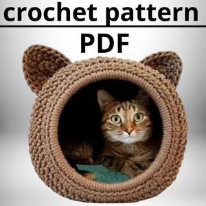 Può includere: Un letto per gatti in crochet di colore marrone con un gatto che sporge dalla apertura. Il letto ha due orecchie in crochet sulla parte superiore. Il testo "crochet pattern PDF" è scritto sopra il letto.