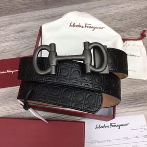 Puede incluir: Cinturón de cuero negro con hebilla color metal de cañón. El cinturón presenta un patrón en relieve repetido y se muestra con una bolsa guardapolvo y una caja roja. La marca "Salvatore Ferragamo" es visible.